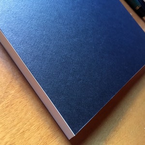 Puede incluir: Un cuaderno azul oscuro con un borde blanco. El cuaderno está sobre una superficie de madera.