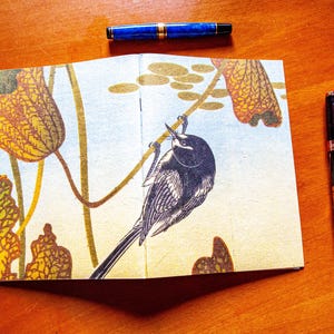 May include: A notebook featuring a print of a black and white bird perched on a stem surrounded by yellow and brown leaves, accompanied by three fountain pens. One blue pen is positioned above the notebook, while two black pens lie to the right.