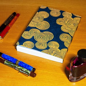 May include: A blue notebook with a gold spiral pattern cover. The notebook is lying on a wooden surface with two pens and a bottle of ink.