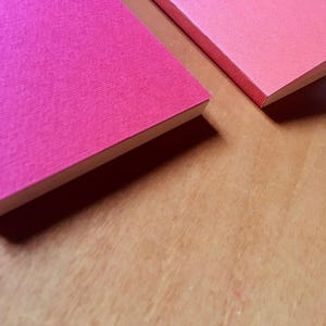 A close-up angled shot shows two pink textured paper notebook covers resting on a wooden surface with a portion of a fountain pen visible.