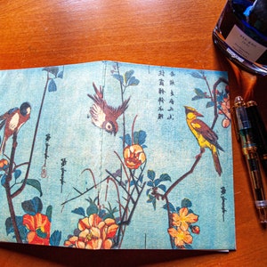 May include: A blue and white Japanese-style notebook with a floral and bird design. The notebook is open to reveal the inside pages, which are blank. The notebook is surrounded by a blue ink bottle and two pens.