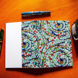 Può includere: Un taccuino aperto con un motivo floreale, con fiori rossi, bianchi e blu su sfondo blu scuro. Tre penne stilografiche e un flacone d'inchiostro sono disposti su una superficie di legno.