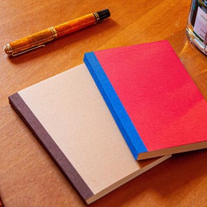 Op de afbeelding: Twee rechthoekige notitieboekjes met een stoffen textuur op de kaft. Het onderste notitieboekje is beige met een donkerbruine rug, terwijl het bovenste notitieboekje felrood is met een felblauwe rug. Drie vulpennen en een inktpotje.