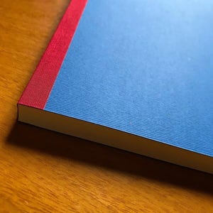 May include: A rectangular notebook featuring a solid blue cover with a textured finish. The spine of the notebook is accented with a vertical strip of red fabric, providing a distinct contrast to the blue cover. The edges are clean and sharp.