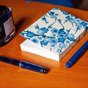 Könnte beinhalten: Eine blaue Tintenflasche, ein blauer Füllfederhalter und ein kleines Notizbuch mit weißem Hintergrund und blauem Blumenmuster.