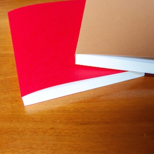 Pode incluir: Dois cadernos retangulares com capas lisas, um em vermelho brilhante e outro em castanho, estão posicionados ao lado de duas canetas-tinteiro. Os cadernos têm páginas brancas visíveis nas bordas e as canetas têm corpos marmoreados em preto e laranja.