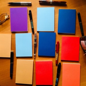 May include: A collection of rectangular notebooks in purple, light blue, dark blue, orange, and red, arranged with various fountain pens, a pair of gold-rimmed glasses, and a glass ink bottle. The items are spread out on a smooth wooden surface.
