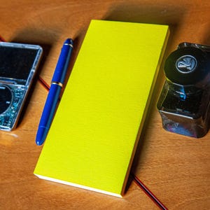 May include: A silver digital music player with a black circular click wheel, a blue fountain pen with gold accents, a bright yellow rectangular notepad, and a clear glass ink bottle with a black cap featuring a swan logo.