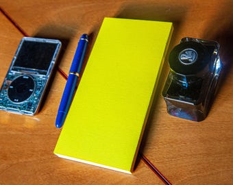 May include: A silver digital music player with a black circular click wheel, a blue fountain pen with gold accents, a bright yellow rectangular notepad, and a clear glass ink bottle with a black cap featuring a swan logo.