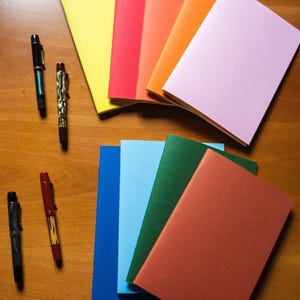 Könnte beinhalten: Eine Sammlung von neun rechteckigen Notizheften mit einfarbigen Papierumschlägen in Gelb, Rot, Orange, Hellrosa, Blau, Hellblau, Grün und Terrakotta, angeordnet in zwei Gruppen neben vier Füllfederhaltern mit unterschiedlichen Designs.