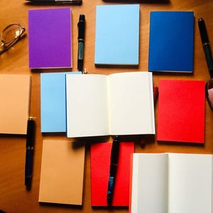 Two passport notebooks are opened to blank white pages among a collection of closed colorful notebooks, fountain pens, glasses, and an ink bottle on wood.
