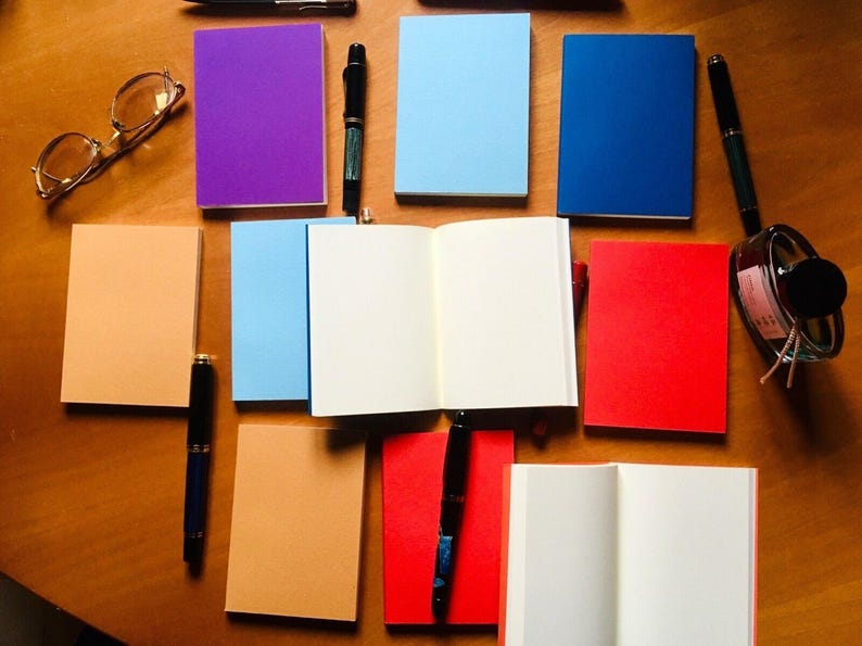 Two passport notebooks are opened to blank white pages among a collection of closed colorful notebooks, fountain pens, glasses, and an ink bottle on wood.