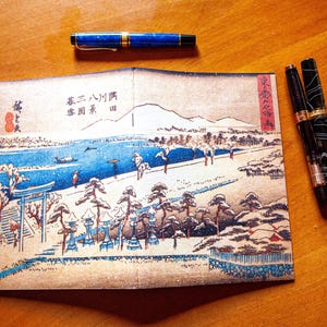 May include: A rectangular print featuring a traditional Japanese landscape illustration with a blue river, pine trees, and a torii gate, accompanied by three fountain pens: one blue with gold trim, one black, and one black with a white marbled pattern.