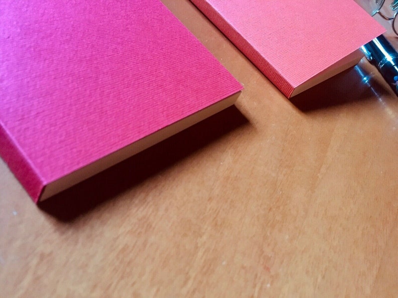 May include: Two rectangular notebooks with textured covers, one in a vibrant magenta and the other in a lighter coral red. The notebooks are positioned side by side, with a black pen visible in the upper right corner of the frame.