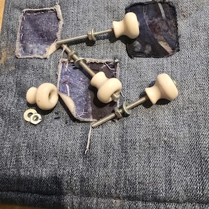 Könnte beinhalten: Ein Jeansstoff mit drei cremefarbenen Keramikknöpfen, die mit Metallschrauben, Unterlegscheiben und Muttern befestigt sind. Der Jeansstoff hat aufgenähte Flicken aus dunklerem blauen Stoff. Die Knöpfe sind für einen Schrank oder eine Schublade.