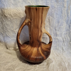 May include: A wooden vase with a green glazed top and two handles. The vase is shaped like a tree trunk with a natural wood grain pattern.