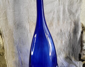 Può includere: Una bottiglia alta in vetro blu cobalto con un design unico e curvo. La bottiglia ha un collo lungo e sottile e una base larga e arrotondata.
