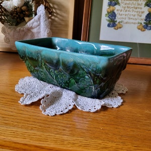 May include: A turquoise and green glazed ceramic planter with a floral design. The planter is sitting on a white crocheted doily.