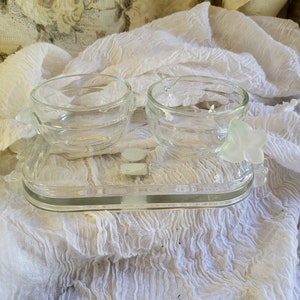 May include: Two clear glass bowls with handles on a clear glass tray. The bowls are empty and the tray is on a white fabric surface.