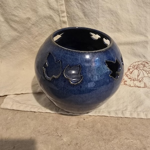 May include: A round, blue ceramic candle holder with leaf-shaped cutouts. The candle holder has a glossy finish and a dark blue interior. The cutouts allow light to shine through, creating a decorative effect. The item is sitting on a neutral-colored surface.