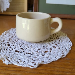 May include: A small, cream-colored ceramic mug with a handle. The mug is sitting on a white crocheted doily.