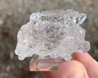 May include: A clear, crystalline cluster of quartz crystals on a small, clear display stand. The crystals are clustered together in a natural, organic shape.