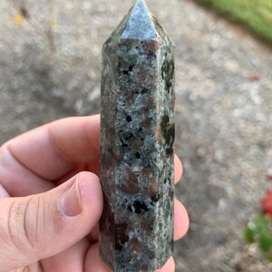 May include: A dark green and black crystal point with a pointed tip and a smooth, polished surface. The crystal has a speckled pattern of black and brown.
