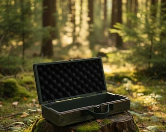 Può includere: Una valigetta verde scuro aperta, con interno in schiuma nera, è appoggiata su un ceppo coperto di muschio in una foresta. La valigetta ha un manico e una forma rettangolare. Lo sfondo mostra alberi alti e luce solare.