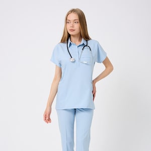 May include: Light blue medical scrubs set, including a short-sleeved top with a pocket and matching pants. A black stethoscope hangs around the neck. The outfit is paired with white sneakers.