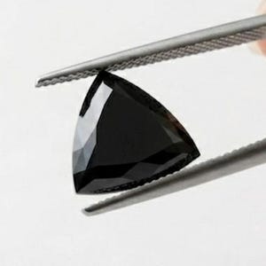 Op de afbeelding: Een gefacetteerde zwarte edelsteen geslepen in een driehoekige vorm. De steen heeft scherpe, strakke randen en een gepolijst oppervlak dat licht reflecteert, wat de diepe, donkere kleur en geometrische vorm benadrukt, vastgehouden door een metalen pincet.