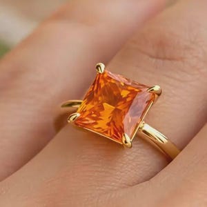 May include: A gold ring featuring a large, square-cut, vibrant orange gemstone held in place by four prongs. The band is smooth and polished, showcasing a simple design that highlights the faceted, brilliant orange stone at the center of the piece.