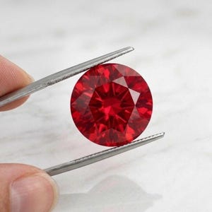 Può includere: Una gemma rossa sfaccettata, rotonda e taglio brillante è tenuta da una pinzetta. La gemma è di un colore rosso intenso e ricco e riflette la luce, mostrando le sue molteplici sfaccettature. La pinzetta è color argento.