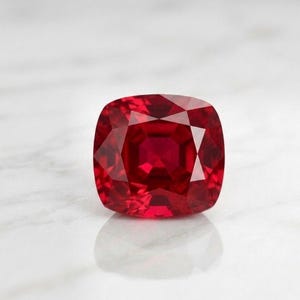 May include: A square-shaped, faceted ruby gemstone. The gem is a deep, vibrant red colour with multiple facets that reflect light. The gemstone is set against a white background, with its reflection visible below.