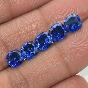May include: Five cushion-cut blue sapphires arranged in a linear row. Each gemstone features a faceted surface that reflects light, displaying a deep, saturated blue color with high clarity and a brilliant, polished finish across all five stones.