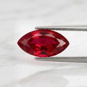 May include: A marquise-cut ruby gemstone, held by tweezers. The ruby is a deep red colour, with multiple facets reflecting light. The gemstone is set against a white background. The ruby is held by silver tweezers.