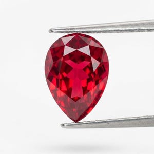Può includere: Una gemma sfaccettata a forma di pera, di colore rosso intenso, tenuta da pinzette d'argento. Il taglio della gemma rivela molteplici sfaccettature, creando una brillante esposizione di luce e colore. La gemma è vibrante.