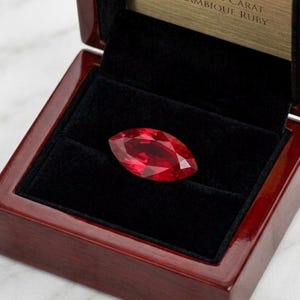 May include: A marquise-cut, vibrant red ruby gemstone displayed in an open, polished wooden jewelry box with a black velvet interior. The ruby's facets reflect light, showcasing its brilliance. The box is a rich, dark red color.