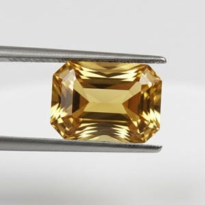 Op de afbeelding: Een goudgele, rechthoekig geslepen edelsteen, vastgehouden door een zilveren pincet. De facetten van de edelsteen vangen het licht. De pincet houdt de steen tegen een effen witte achtergrond, wat de kleur en slijpvorm benadrukt.