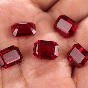 Peut inclure: Cinq pierres précieuses en vrac, de forme rectangulaire et taille émeraude, d'une couleur rouge profond et vibrante. Chaque pierre présente des surfaces facettées qui reflètent la lumière, créant un aspect clair et transparent.