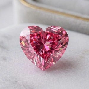 May include: A heart-shaped, faceted pink gemstone. The gem has a deep pink center that fades to a lighter pink towards the edges. The stone is set against a white surface and a gray jewelry box.