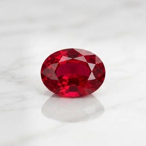 May include: A faceted, oval-shaped ruby gemstone. The gem is a deep, vibrant red color, with multiple facets that catch and reflect light. The ruby is set against a white background, with a subtle shadow.