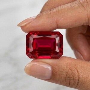 Può includere: Una gemma rosso scuro di grandi dimensioni, tagliata a forma rettangolare. La gemma è tenuta tra due dita, mostrando le sue sfaccettature e la sua brillantezza interna. Il colore della pietra è un rosso ricco e saturo.