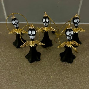 May include: Five black and white skull angel ornaments with gold wings and halos. The ornaments are hanging from gold cords.