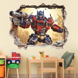 May include: A wall decal featuring a 3D-style illustration of Optimus Prime in red and blue armor, holding a large blaster. The character appears to be breaking through a brick wall, with debris scattered around the edges of the rectangular frame.