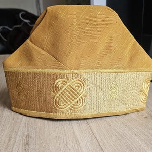 May include: A gold-colored hat with a structured crown and a decorative band. The band features embroidered geometric patterns and scrollwork. The hat appears to be made of a textured fabric.