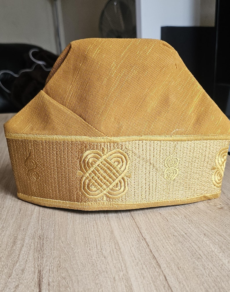 May include: A gold-colored hat with a structured crown and a decorative band. The band features embroidered geometric patterns and scrollwork. The hat appears to be made of a textured fabric.