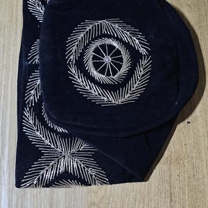 May include: Two black velvet fabric squares with silver embroidery. The embroidery is a circular design with a smaller circle in the center.