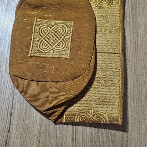 May include: A gold-colored prayer cap and mat set. The cap is oval-shaped with an embroidered square design. The mat is rectangular with a matching embroidered pattern. Both items have a textured appearance and are made of a shiny, gold fabric.