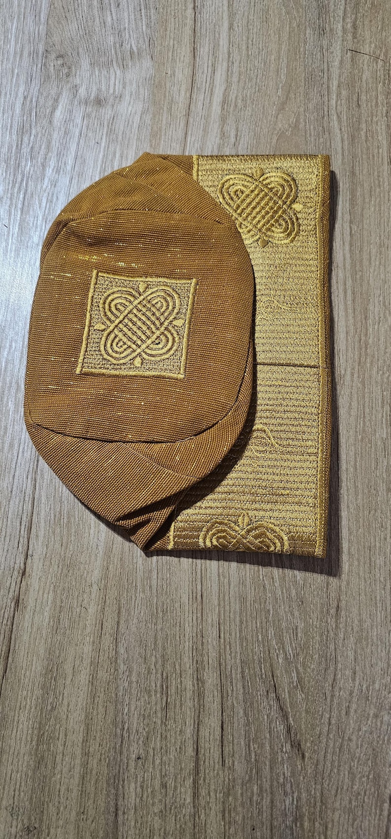 May include: A gold-colored prayer cap and mat set. The cap is oval-shaped with an embroidered square design. The mat is rectangular with a matching embroidered pattern. Both items have a textured appearance and are made of a shiny, gold fabric.
