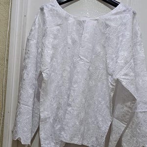 Puede incluir: Una blusa blanca de manga larga con un intrincado patrón de encaje floral. La blusa está colgada de una percha negra. Las mangas tienen un diseño acampanado y la tela parece ser semi-transparente.